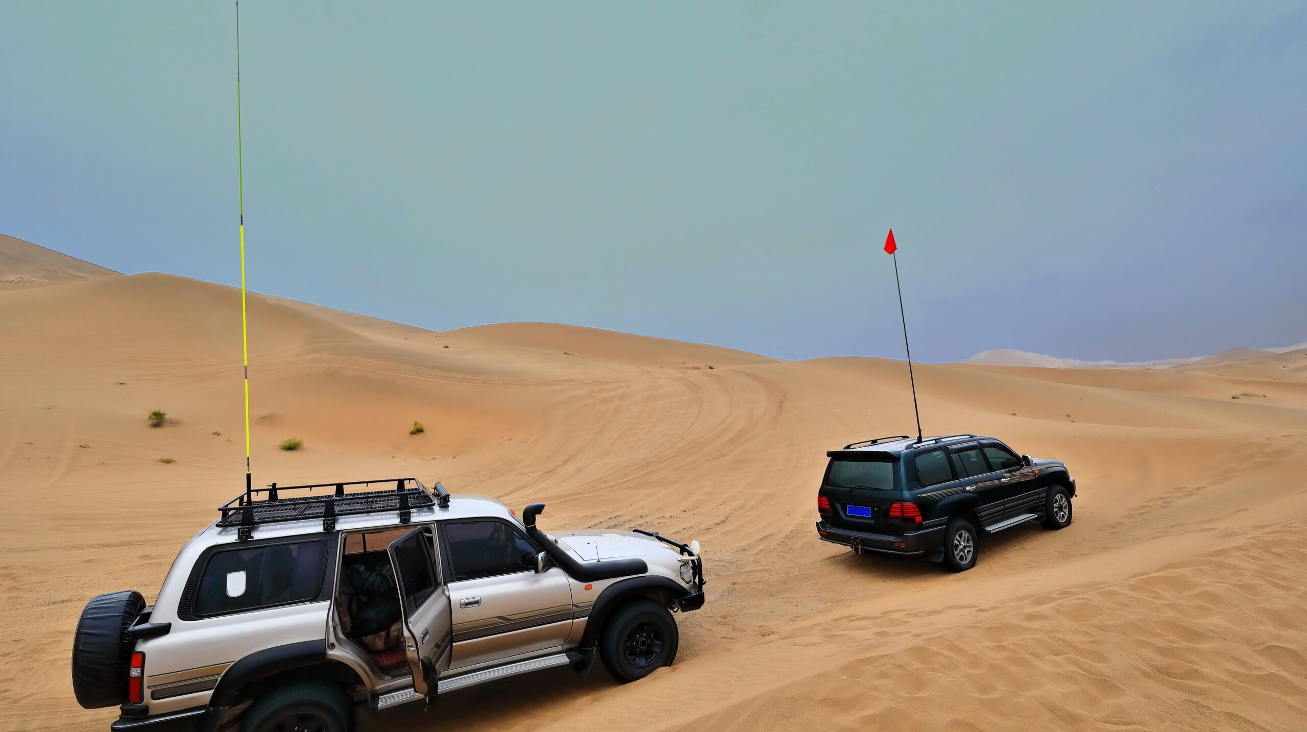 Gray green tourist off-road cars in the sand-long flagpoles and pennants to be seen among the megadunes. Badain Jaran-Badanjilin Shamo-Mysterious Lakes Desert-sector of Gobi Desert-InnerMongolia-China