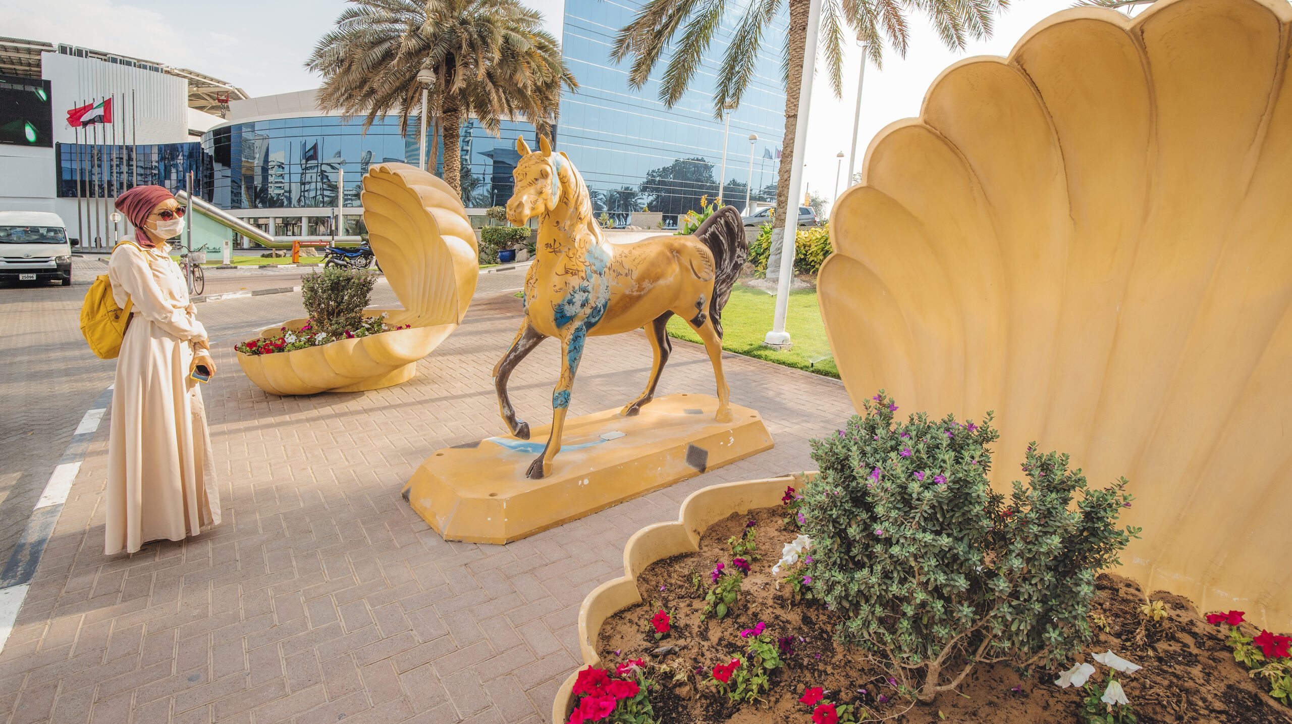 23 February 2021, Dubai, UAE: Woman in a medical mask walks along a city street during a coronavirus quarantine and admires the sculpture of a mythical horse