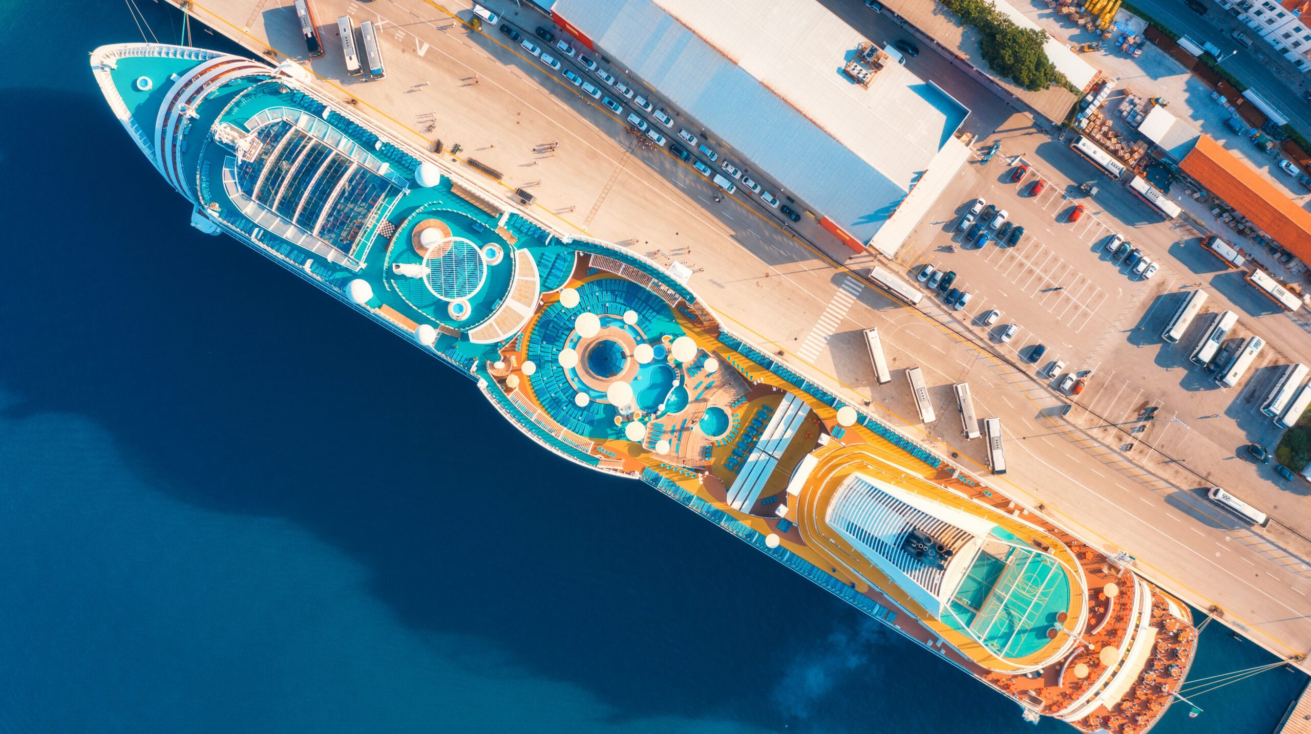 Aerial view of luxury cruise liner in port. Top view of swimming pool with sunbeds, umbrellas and deck chairs, wooden deck on the cruise ship, cars and buildings in summer. View from above. Resort