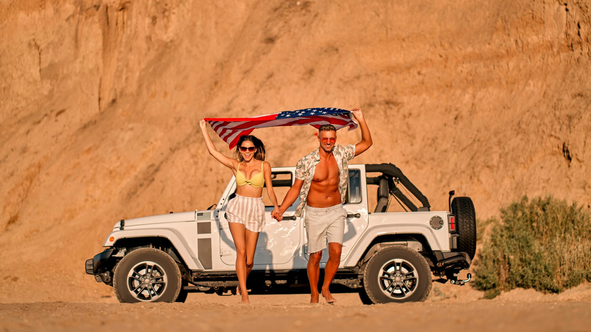 Romantic couple with car parked on the beach. Summer vibes. Traveling together to the sea. Handsome man and beautiful woman walking with American flag in hands