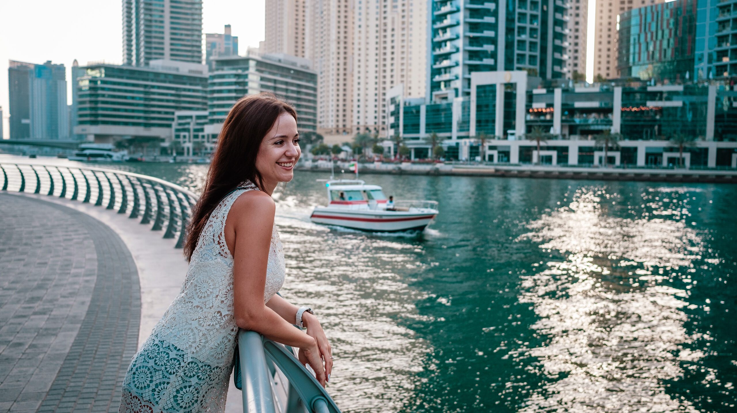 Enjoying travel in United Arabian Emirates. Happy young woman. Amazing view of sea and city. Girl smiles to people sailing on a yacht and waves her hand