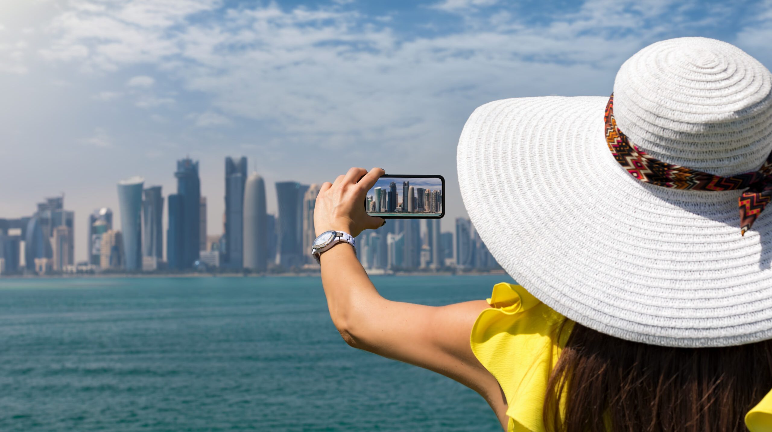 rear-view-woman-photographing-cityscape-with-smart-phone-by-sea-against-sky
