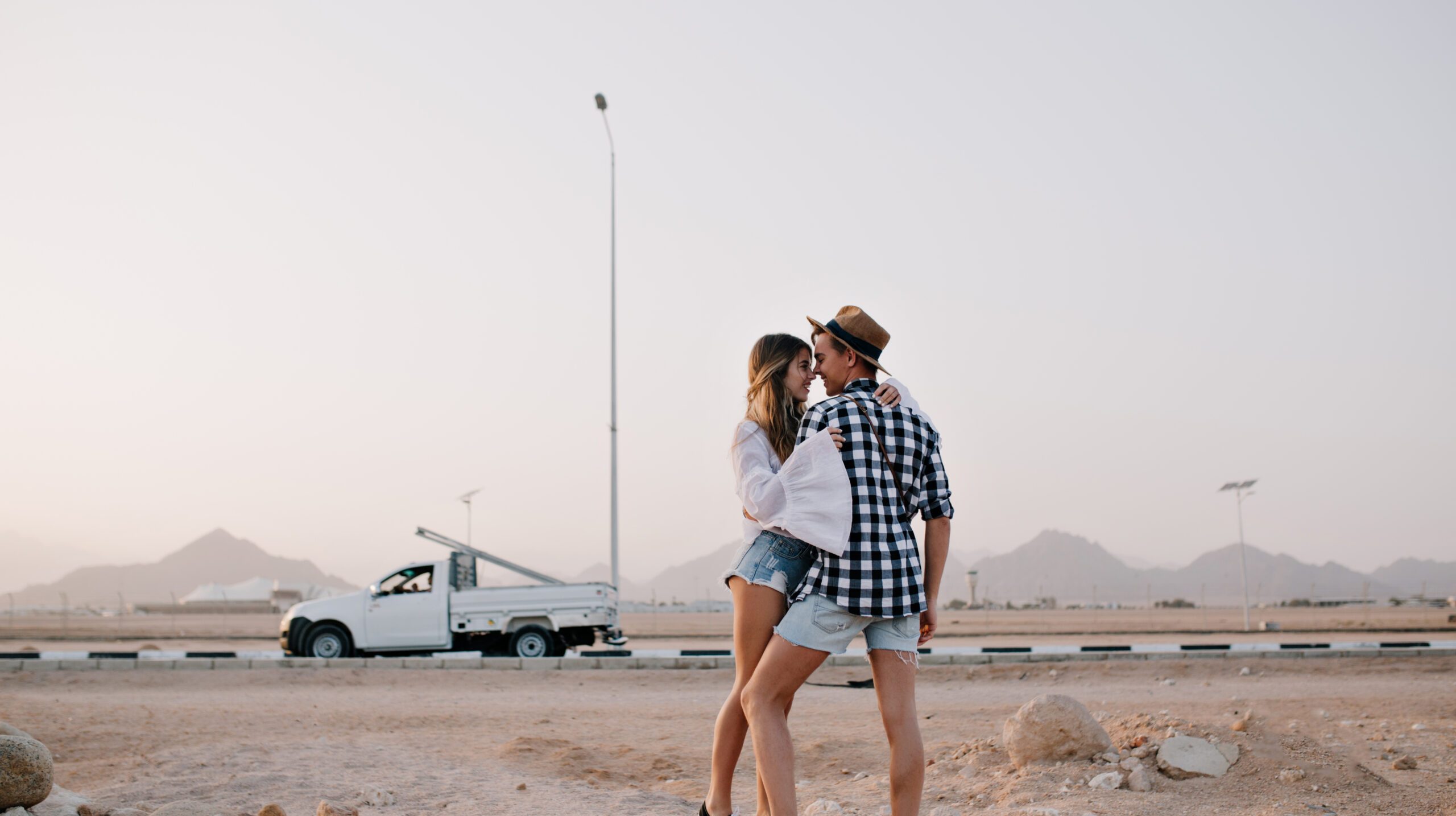 Slender pretty girl in vintage white shirt wants to kiss her boyfriend, standing on tiptoe under gray sky. Beautiful loving couple hugging in front of car and enjoys amazing nature views in evening
