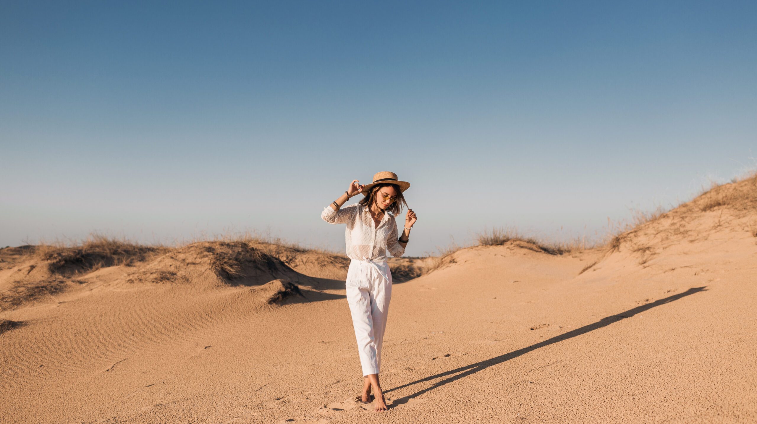 stylish beautiful woman walking in desert sand in white outfit wearing straw hat on sunset, travel safari on vacation