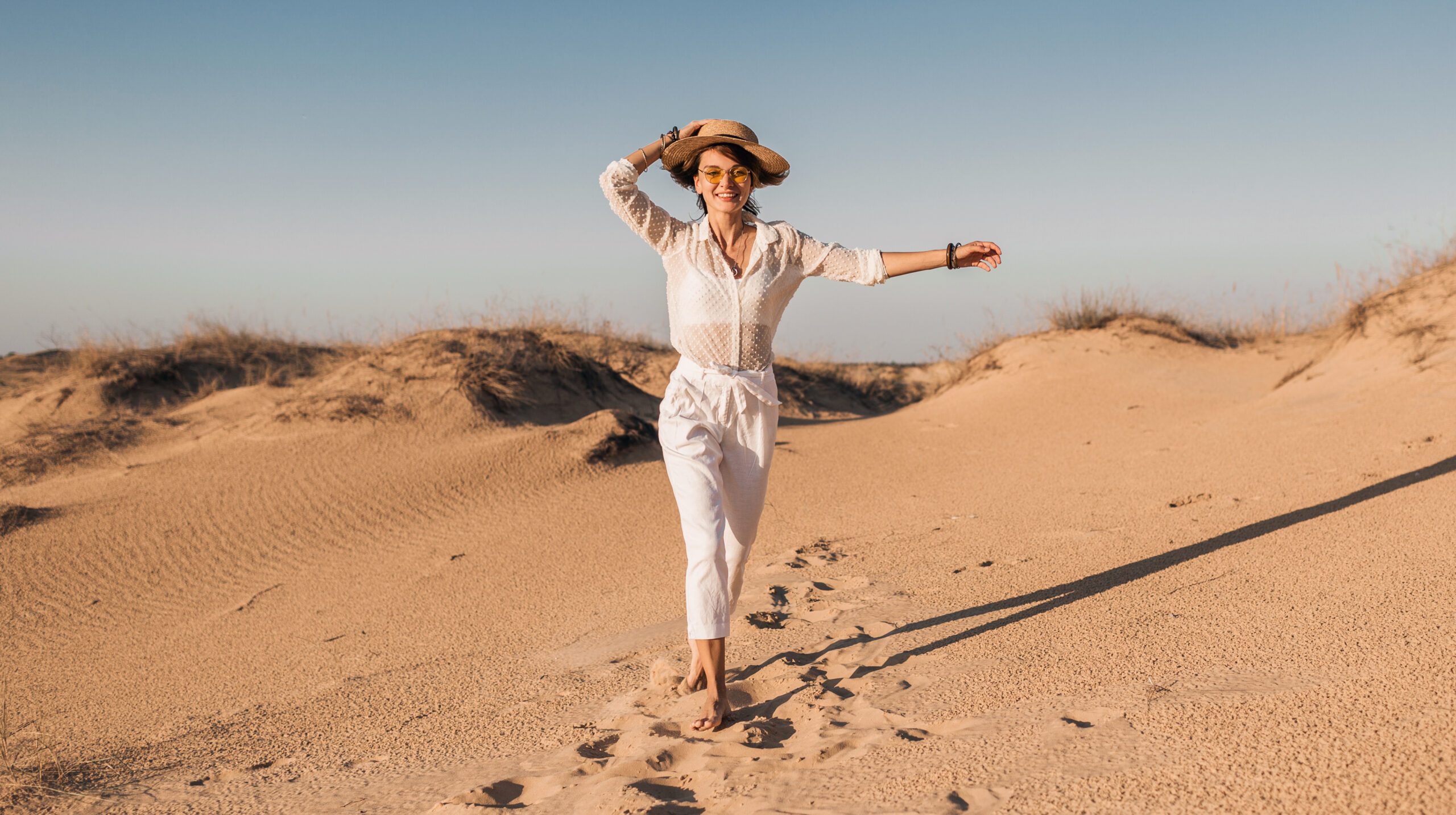 stylish smiling beautiful happy woman running and jumping in desert sand in white outfit wearing straw hat on sunset, travel safari on vacation