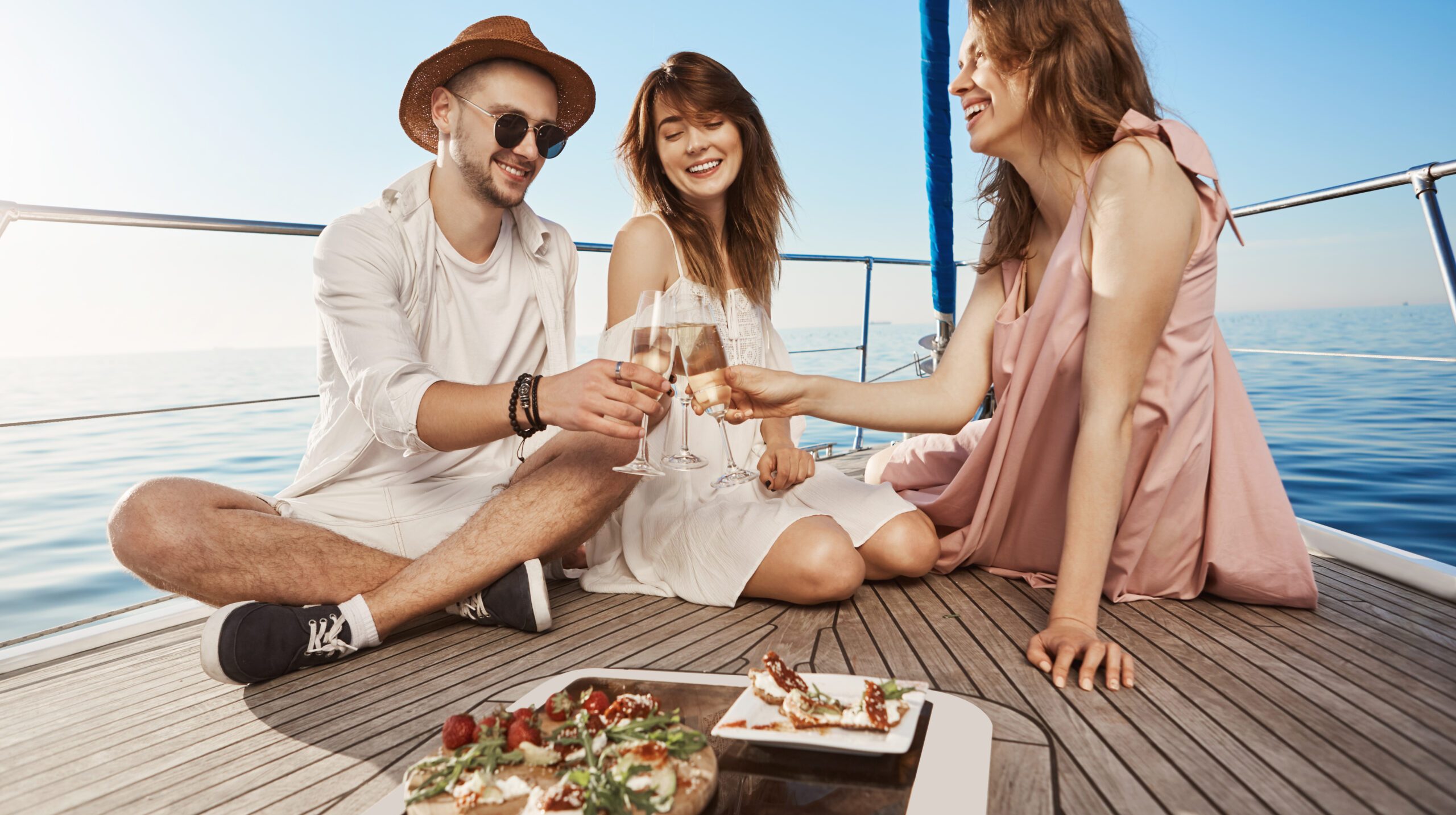 Outdoor shot of three trendy european friends sitting on boat, having lunch and drinking champagne, expressing joy and pleasure. Every year they book tickets to warm countries in winter. Copy space.