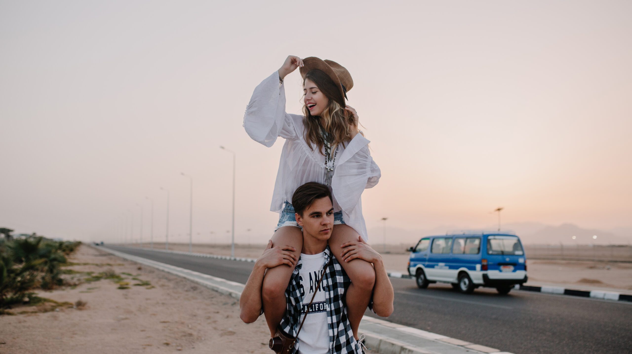 Tired brunette guy in plaid shirt with a retro camera carries his girlfriend on his shoulders looking at sunset. Cheerful young woman in trendy hat having fun with boyfriend on outdoor evening date