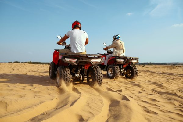 Two men in helmets riding on atv in desert sands, back view. Male persons on quad bikes, sandy race, dune safari in hot sunny day, 4x4 extreme adventure, quad-biking