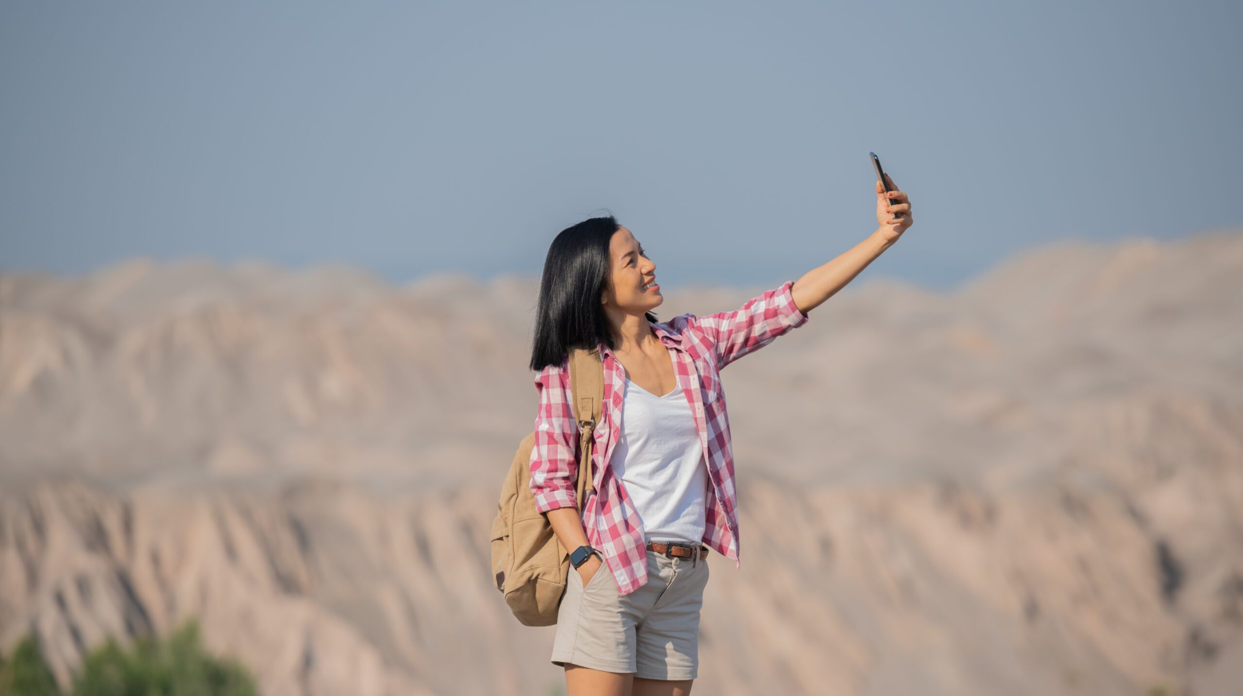 woman hiking in mountains standing on rocky summit ridge with backpack and pole looking out over landscape, happy female making self portrait in mountains, holding smartphone camera to take picture.
