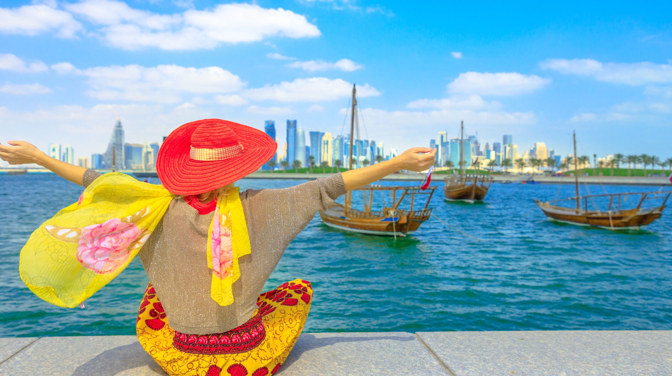 Happy woman on seafront of Doha park along Doha Bay with traditional dhow on background. Lifestyle caucasian tourist enjoys East Mound-Skyline view skyscrapers of Doha Downtown. Qatar, Middle East.