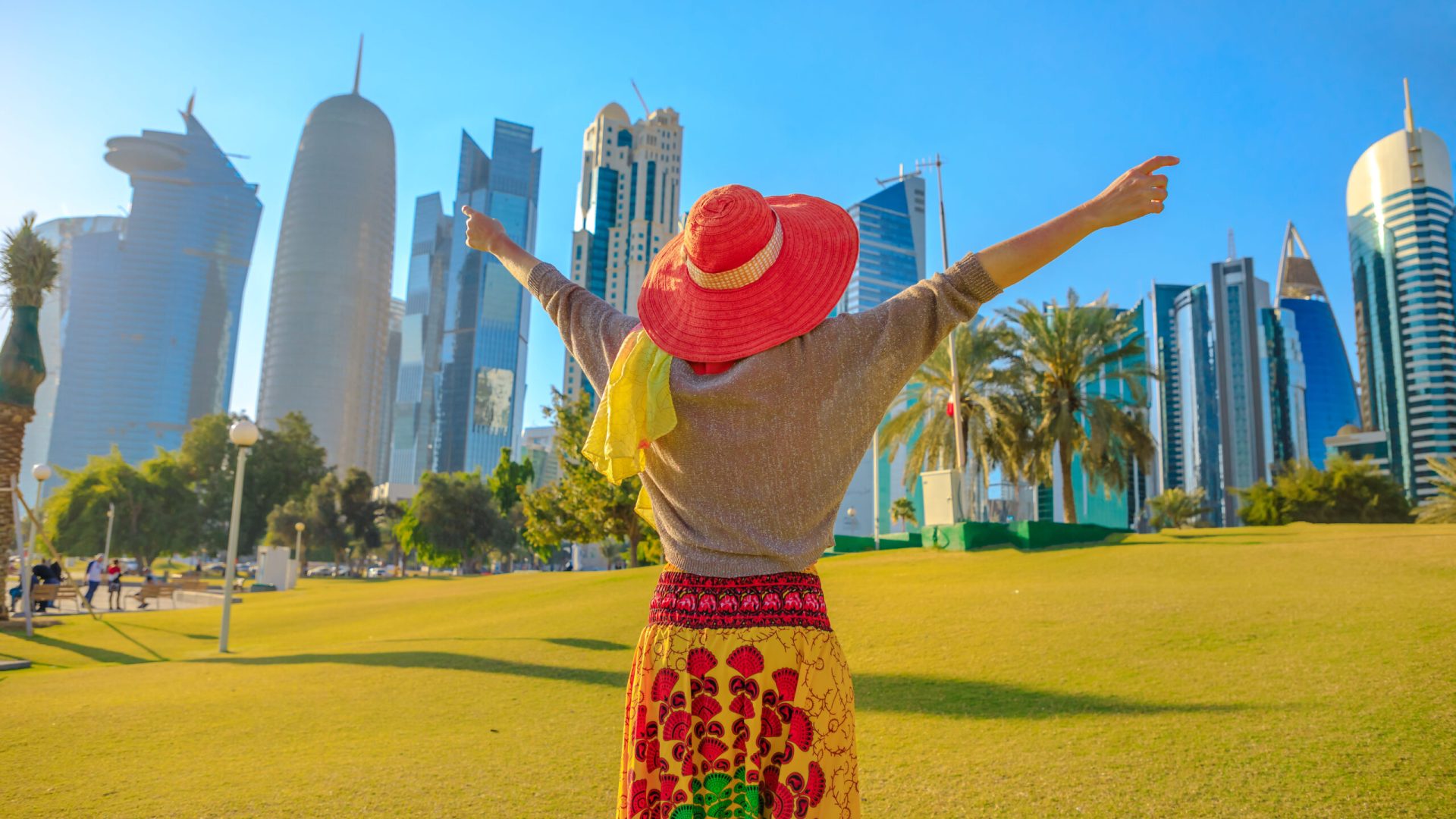 Travel in Qatar. Happy woman with sunhat in a park along Corniche promenade at sunset. West Bay skyline on background. Tourist enjoys in Doha Downtown. Middle East, Arabian Peninsula in Arabian Gulf.