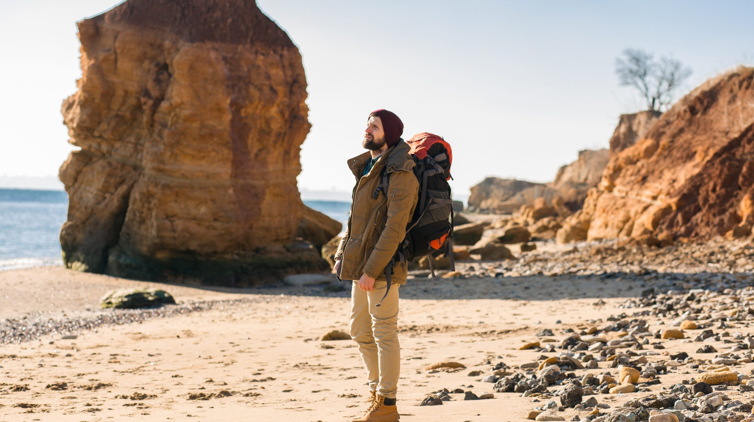 young hipster man traveling alone with backpack in autumn sea coast wearing warm jacket and hat, active tourist, exploring nature in cold season
