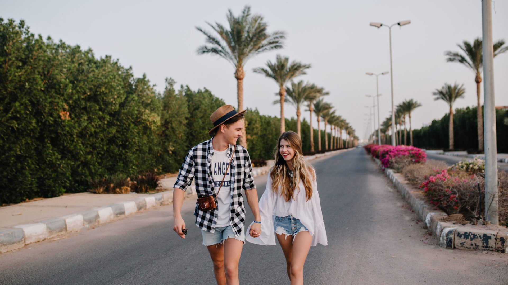 Young man in trendy hat and denim shorts holding girlfriend's hand, walking down the street with bushes on the side. Beautiful couple in stylish outfit spending time outside, enjoys exotic views
